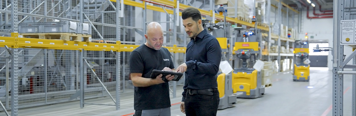 Zwei Männer in Arbeitskleidung stehen im Hochregallager und blicken auf ein Tablet.