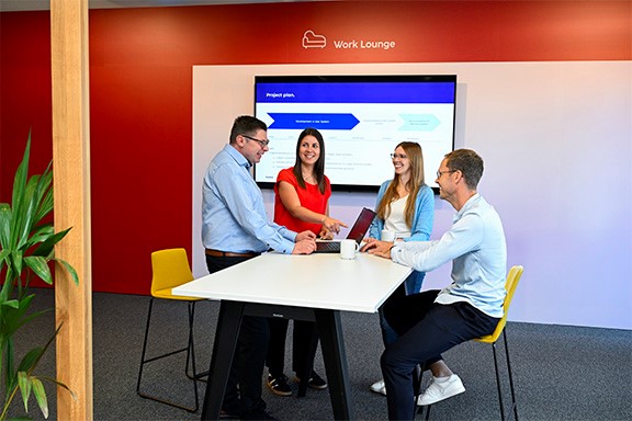 Vier Kollegen stehen im Meetingraum an einem Tisch und unterhalten sich.