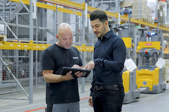 Zwei Männer in Arbeitskleidung stehen im Hochregallager und blicken auf ein Tablet.