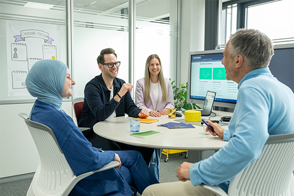Drei Kollegen in einem Büro. Eine Frau betritt mit Dokumenten, der Mann trägt einen Rucksack, die zweite Frau hält Laptop und Headset.